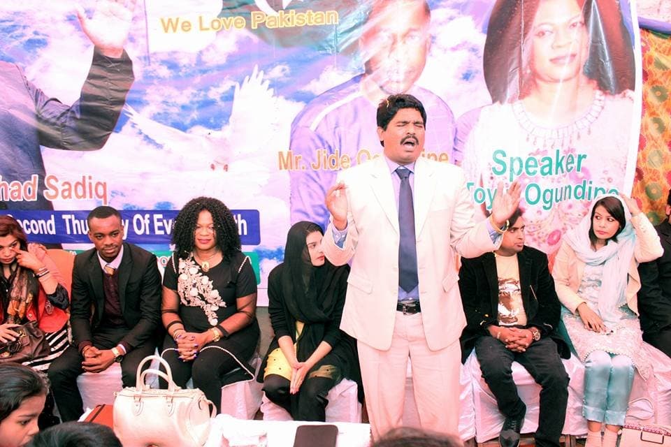 Pastor Arshad Sadiq praying on stage with guests seated behind him
