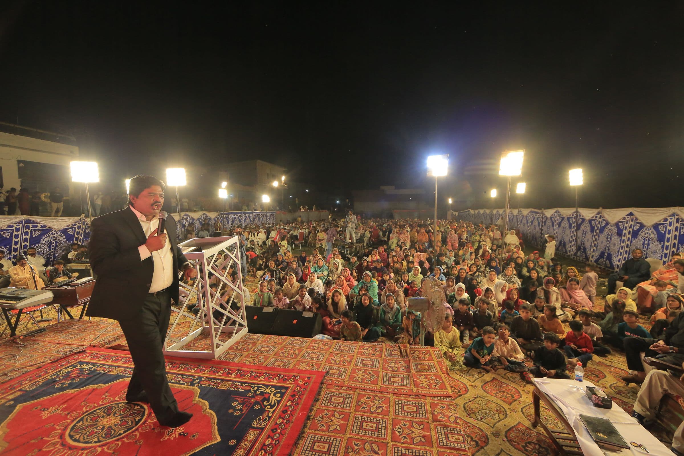 Pastor Arshad Sadiq preaching on stage to a large open-air gathering at night