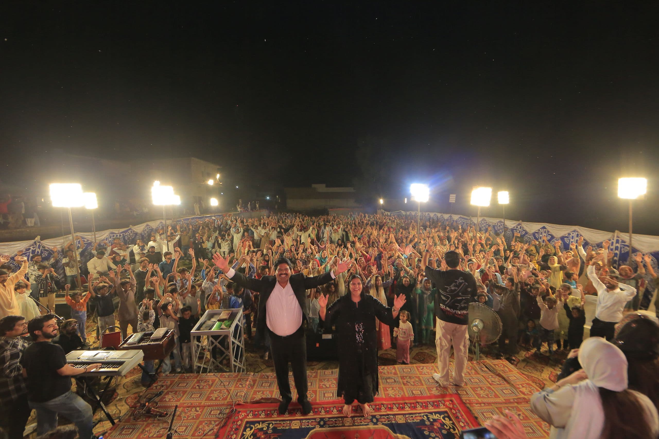 Pastor Arshad Sadiq and leaders on stage as a large crowd lifts their hands at an open-air night service